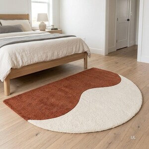 May include: A semi-circular area rug featuring a two-tone design with a textured rust-orange section and a solid cream-colored section. The rug has a soft, plush pile and is positioned on a light-colored wooden floor in a bedroom setting.