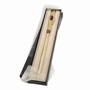 Puede incluir: Una varita larga y delgada con un eje pálido y liso y un mango texturizado de color dorado que presenta una incrustación ovalada de color rojo intenso. La varita se presenta dentro de una caja rectangular negra forrada con tela crema.