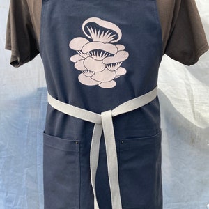May include: A dark blue apron with a beige mushroom graphic. The apron has two front pockets, a beige waist tie, and beige neck straps with metal grommets. The apron is displayed on a mannequin.