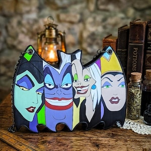 Op de afbeelding: Zwarte handtas in de vorm van een vleermuis, met kleurrijke illustraties van Disney-schurken. De tas heeft de gezichten van Maleficent, Ursula, Cruella de Vil en de boze koningin en een kettingriem.