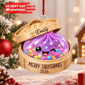 May include: A hanging Christmas ornament shaped like a bamboo steamer basket containing a purple, winking dumpling character filled with colorful spheres and stars. The lid features the name Emily, and the base reads MERRY SQUISHMAS 2026.