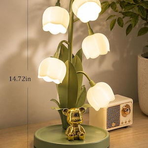 May include: A decorative table lamp shaped like a lily flower, with five illuminated white flower-shaped shades. The lamp is 14.72 inches tall and 7.08 inches wide, with a gold bear figurine on the base. The lamp is green and white.