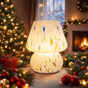 May include: A glowing mushroom-shaped lamp with a white base and shade, adorned with blue and yellow accents. The lamp is set on a table with Christmas decorations, including a red gift box, ornaments, and a Christmas tree in the background.
