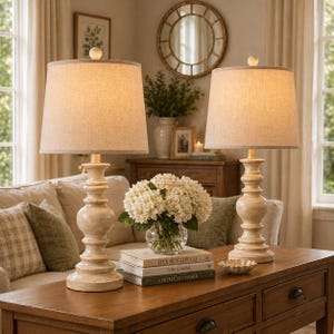 May include: Two cream-colored table lamps with beige shades sit on a wooden table. The lamps have a classic, ornate base design. A vase of white flowers and stacked books are also on the table, creating a cozy, inviting atmosphere.