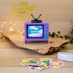 Puede incluir: Un mini televisor táctil morado con el texto "The Simpsons" en la pantalla y el nombre del producto arriba. El televisor tiene una antena negra y botones turquesa. También hay pegatinas de personajes de dibujos animados y un cable blanco.