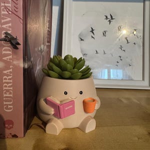 Op de afbeelding: Een grillige, lichtroze keramische plantenpot in de vorm van een karakter, met een roze boek en een oranje mok. De pot is gevuld met een groene vetplant. Een ingelijste foto van vogels in vlucht staat op de achtergrond.
