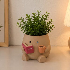 Könnte beinhalten: Ein beigefarbener Keramik-Pflanztopf in Form einer sitzenden Figur mit lächelndem Gesicht, die ein kleines rosa Buch in der einen und eine pfirsichfarbene Tasse in der anderen Hand hält. Der Topf enthält eine grüne Kunstpflanze mit vielen Blättern.