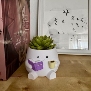Peut inclure: Un pot de plante en céramique blanche en forme de personnage, tenant un livre violet et une tasse de café jaune. Le pot est rempli d'une plante succulente verte. Le pot a un visage souriant et est posé sur une surface en bois.