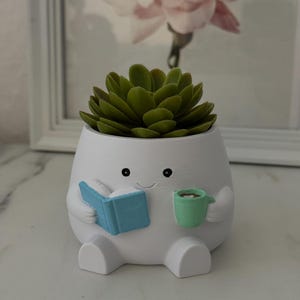 Könnte beinhalten: Ein weißer, figurförmiger Pflanzentopf, der ein blaues Buch und eine grüne Tasse hält und mit einer grünen Sukkulente bepflanzt ist. Die Figur hat ein lächelndes Gesicht. Ein dekorativer Artikel für die Inneneinrichtung.