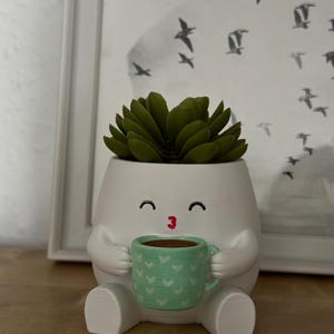 Könnte beinhalten: Ein weißer, eiförmiger Übertopf mit einem Cartoon-Gesicht, geschlossenen Augen und einem kleinen roten Mund. Der Übertopf hat Arme, die eine hellgrüne Tasse mit weißem Herzmuster halten. Eine grüne Sukkulente befindet sich im oberen Teil des Topfes.
