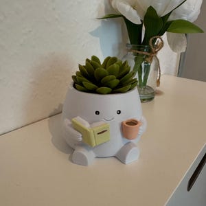 Könnte beinhalten: Ein weißer Keramik-Blumentopf in Form einer Zeichentrickfigur, die ein kleines Buch und eine Tasse hält. Der Topf ist mit einer grünen Sukkulente gefüllt. Im Hintergrund befindet sich eine kleine Glasvase mit weißen Blumen.