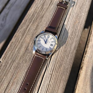 May include: A silver wristwatch with a light-colored face and a brown leather band. The watch has a fluted bezel and a date window at the 3 o'clock position. The brown leather band is stitched with white thread. The watch is displayed on a wooden surface.