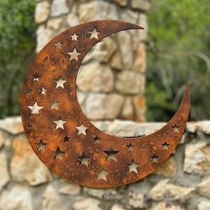 May include: A crescent moon wall art piece crafted from metal with a weathered, rusted brown finish. The surface features numerous star-shaped cutouts of varying sizes distributed across the curved form, creating a celestial decorative design.