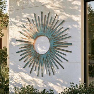 May include: A circular sunburst wall sculpture featuring a weathered teal and copper patina finish. The design consists of a central open ring surrounded by numerous radiating metal spikes of varying lengths, creating a star-like pattern.