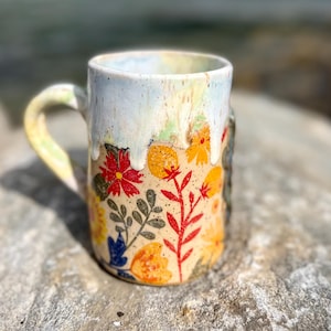 Peut inclure: Une tasse en céramique avec un sommet émaillé blanc qui coule sur une base beige. La partie inférieure présente un motif peint à la main de fleurs rouges, orange, jaunes et bleues avec des feuilles vertes. La tasse a une anse incurvée.