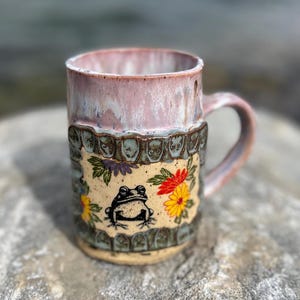Può includere: Una tazza in ceramica con bordo e manico smaltati in rosa e bianco. Il corpo presenta un pannello centrale color crema con un disegno a linee nere di una rana circondato da fiori dipinti in rosso, arancione e giallo, incorniciato da un bordo.