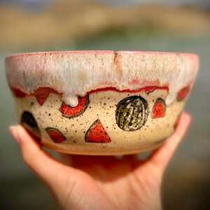 May include: A ceramic bowl with a speckled cream base featuring hand-painted watermelon slices and whole watermelons in red, green, and black. The rim has a thick, dripping white glaze over a pink band, creating a textured, layered appearance.
