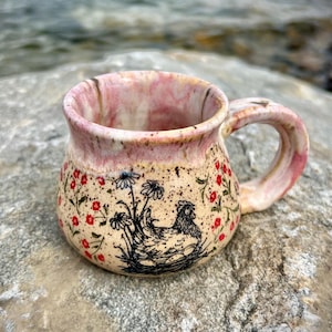 Peut inclure: Une tasse en céramique de forme arrondie, dotée d'une base crème tachetée et d'un bord émaillé rose. L'extérieur est orné d'une illustration au trait noir représentant une poule assise dans un nid entourée de petites fleurs rouges et marguerites.
