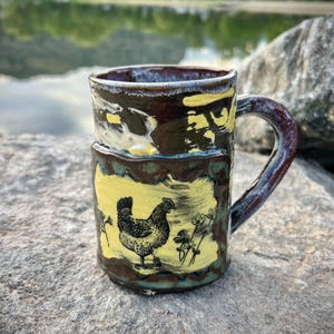 Puede incluir: Taza de cerámica hecha a mano con una ilustración de un pollo. La taza presenta un esmalte marrón y amarillo con un dibujo en blanco y negro de una gallina y flores. El asa y el borde son de color azul oscuro y marrón.