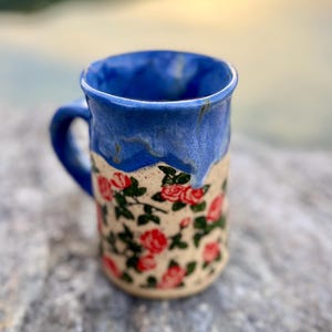 May include: A ceramic mug featuring a speckled cream-coloured base decorated with a pattern of red roses and green leaves. The upper portion of the mug is finished with a vibrant blue glaze that drips down over the floral design, creating a contrast.