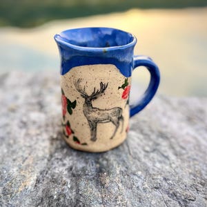 Peut inclure: Une tasse en céramique avec un corps crème moucheté et un bord ainsi qu'une anse émaillés d'un bleu vif. La tasse présente une illustration détaillée à l'encre noire d'un cerf aux grands bois, entouré de petits motifs floraux rouges.
