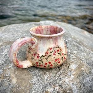 May include: A ceramic mug with a rounded body and a curved handle, featuring a speckled cream base decorated with small red flowers and green stems, topped with a glossy pink and white drip glaze that flows down from the rim.