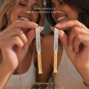 May include: Two women holding matching rectangular pendants. The pendants are silver and gold, with the words "happiness is... laughing at nothing together" engraved on them. The image also includes the text "SOME MESSAGES ARE JUST BETWEEN YOU TWO."