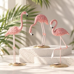 Puede incluir: Tres figuras de flamencos en diversas posturas, cada una con un cuerpo rosa texturizado, patas largas y delgadas, y un pico con punta negra. Las figuras están montadas sobre bases circulares de metal dorado, mostrando distintas posiciones.