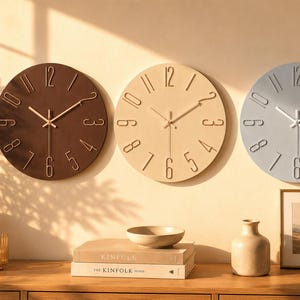 May include: Three circular wall clocks arranged horizontally, each featuring raised numerals and thin hands. The left clock is dark brown, the centre clock is beige, and the right clock is light grey. Each clock has a smooth, matte finish.