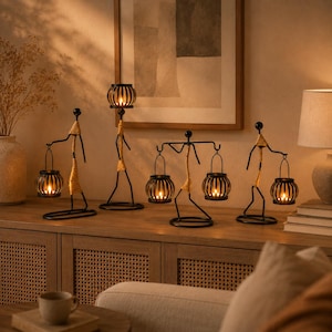 May include: A set of four black metal candle holders shaped like stylised figures with wrapped tan fibre accents. Each figure supports one or two spherical, slatted metal lanterns containing lit candles, creating a warm, glowing display.