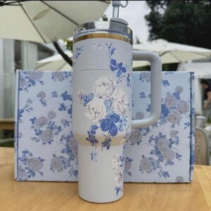 Op de afbeelding: Een lichtgrijze ge&iuml;soleerde beker met een handvat, met een bloemenpatroon van witte en blauwe rozen. De beker heeft een zilveren deksel met een rietje en een gouden accent. Een bijpassende doos met hetzelfde bloemenpatroon staat achter de beker.