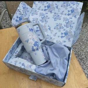 Op de afbeelding: Een lichtblauwe ge&iuml;soleerde beker met een handvat, versierd met witte en blauwe bloempatronen, gepresenteerd in een bijpassende bloempatroon doos. De beker heeft een gouden rand en een doorzichtige rietje. Een lichtblauwe doek zit ook in de doos.