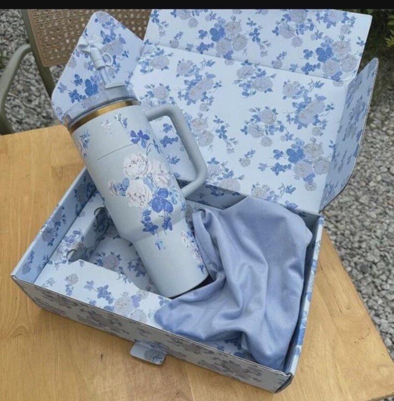 Op de afbeelding: Een lichtblauwe ge&iuml;soleerde beker met een handvat, versierd met witte en blauwe bloempatronen, gepresenteerd in een bijpassende bloempatroon doos. De beker heeft een gouden rand en een doorzichtige rietje. Een lichtblauwe doek zit ook in de doos.