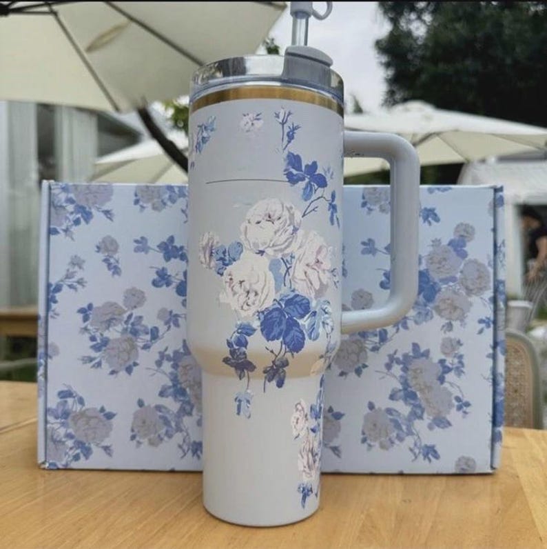 Op de afbeelding: Een lichtgrijze ge&iuml;soleerde beker met een handvat, versierd met een bloemenpatroon in wit en blauw. De beker heeft een zilveren deksel met een rietje en een gouden accent. Een bijpassende doos met hetzelfde bloemenpatroon staat achter de beker.