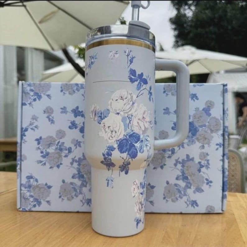 Op de afbeelding: Een lichtgrijze ge&iuml;soleerde beker met een handvat, met een bloemenpatroon van witte en blauwe rozen. De beker heeft een zilveren deksel met een rietje en een gouden accent. Een bijpassende doos met hetzelfde bloemenpatroon staat achter de beker.