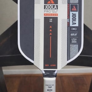 May include: A Joola Pro Perseus 16 mm table tennis paddle. The paddle has a black and white design with the text "JOOLA PRO IV PERSEUS" and "BEN JOHNS". The handle has the text "REMOVE FOR TAP HERE".