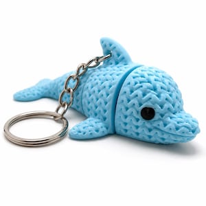 May include: A light blue dolphin keyring with a textured surface. The dolphin has a silver chain and keyring attached. The dolphin's eye is black. The keyring is on a white background.