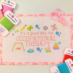 May include: A needlepoint canvas featuring the text It's a good day for OCCUPATIONAL THERAPY surrounded by colorful stitched icons including a pencil, swing, teddy bear, and stacking toy, accompanied by pink scissors and Tilli Tomas silk thread.