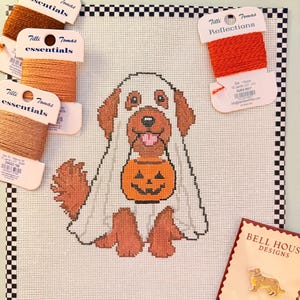 May include: A needlepoint canvas featuring a brown dog wearing a white ghost costume and holding an orange jack-o'-lantern bucket. The canvas is surrounded by three spools of Tilli Tomas Essentials thread, one spool of Tilli Tomas Reflections thread, and a Bell House Designs pin.