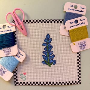 May include: A square needlepoint canvas featuring a blue lupine flower design and a small heart, bordered by a black and white checkerboard pattern. Included are pink embroidery scissors and four Tilli Tomas silk thread cards in blue and green.