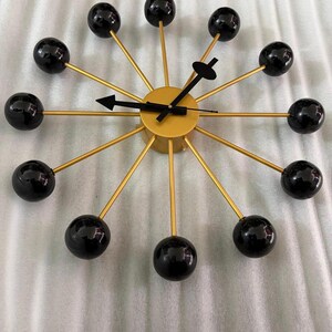 May include: A modern wall clock with a gold-colored face and arms. The clock features black spheres at the end of each arm, and black clock hands. The clock is a decorative piece for any room.