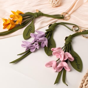 Peut inclure: Trois porte-clés en cuir en forme de fleurs jaunes, lilas et roses. Chaque porte-clés présente un bouquet de fleurs avec des feuilles vertes et un anneau doré. Les feuilles sont gravées des noms : Olivia et Amelia.