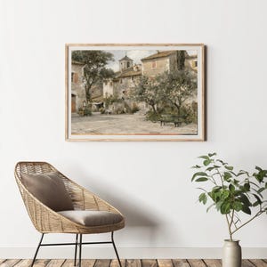 May include: A framed print of a European village square, featuring stone buildings, trees, and a bench. The artwork is in a muted color palette, with a light brown wooden frame. The image is displayed above a wicker chair and a potted plant.