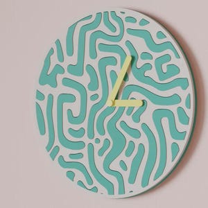 May include: A round wall clock with a white face featuring a teal abstract pattern. The clock hands are yellow. The clock is mounted on a light pink wall.