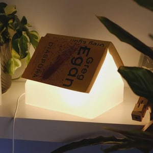 Op de afbeelding: Een wit, huisvormige lamp die licht geeft, met een boek getiteld "Diaspora" van Greg Egan erop. De lamp staat op een witte plank, met een plant en een cilindrisch object aan de linkerkant.