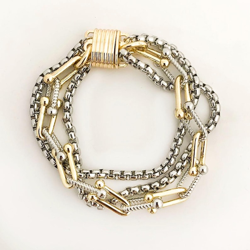 K&ouml;nnte beinhalten: Mehrstrang-Armband aus silber- und goldfarbenem Metall. Das Armband hat verschiedene Kettenarten, darunter Boxketten und U-f&ouml;rmige Glieder. Der Verschluss ist goldfarben und zylindrisch.