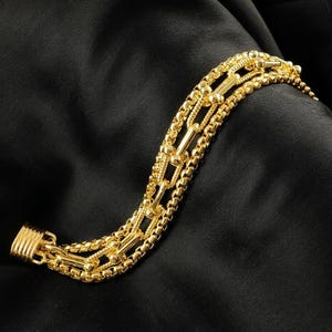 K&ouml;nnte beinhalten: Ein goldfarbenes Armband mit mehreren Str&auml;ngen verschiedener Kettenarten. Das Armband hat eine Mischung aus Box-, Perlen- und Stabgliedern, die einen geschichteten Effekt erzeugen. Der Verschluss ist einfach und sicher. Der Schmuck wird vor schwarzem Hintergrund gezeigt.