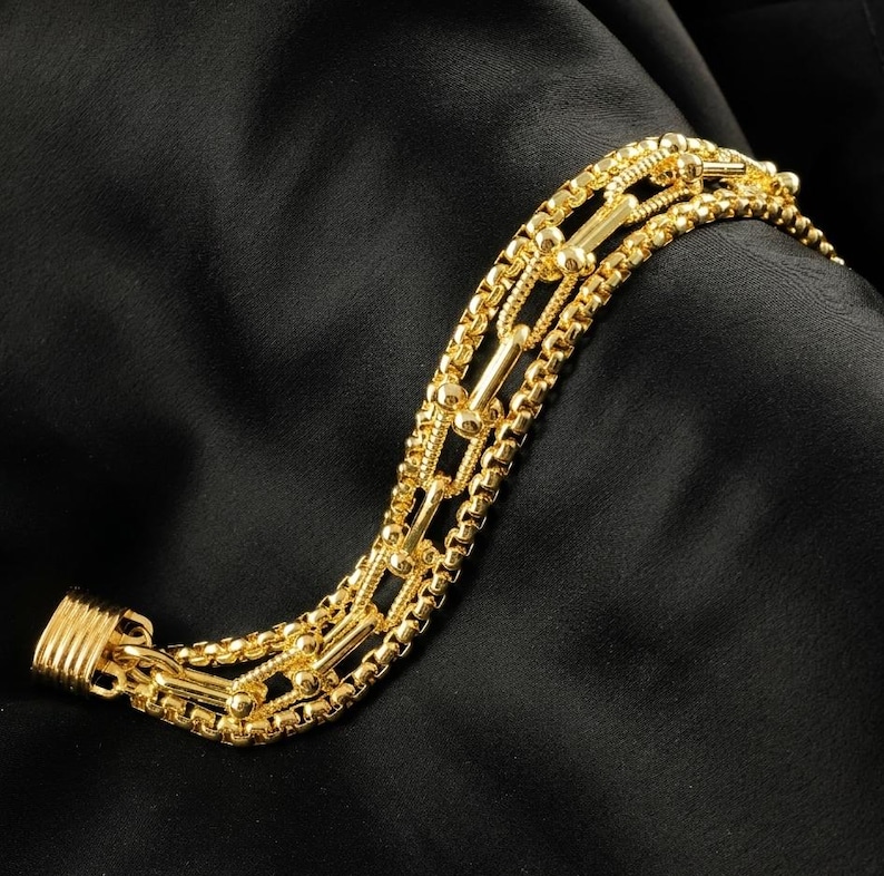 K&ouml;nnte beinhalten: Ein goldfarbenes Armband mit mehreren Str&auml;ngen verschiedener Kettenarten. Das Armband hat eine Mischung aus Box-, Perlen- und Stabgliedern, die einen geschichteten Effekt erzeugen. Der Verschluss ist einfach und sicher. Der Schmuck wird vor schwarzem Hintergrund gezeigt.