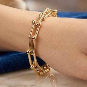 Puede incluir: Un brazalete dorado con un diseño de eslabones de cadena. El brazalete está hecho de eslabones en forma de U conectados con pequeños acentos redondos. El brazalete se usa en una muñeca y los eslabones son visibles.