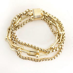 K&ouml;nnte beinhalten: Ein goldfarbenes Armband mit mehreren Str&auml;ngen verschiedener Kettenarten. Das Armband umfasst Boxketten, Kugelkettchen und einen Magnetverschluss. Der Schmuck wird auf wei&szlig;em Hintergrund pr&auml;sentiert.